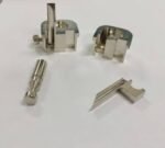 Assorted small silver metal mechanical components on a white surface, including clamps, a pin-like piece, and a wedge-shaped part.