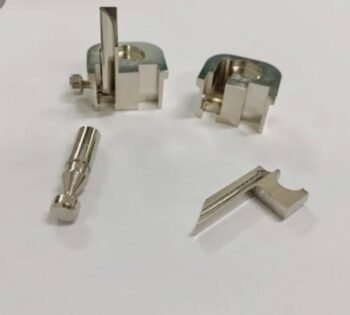 Assorted small silver metal mechanical components on a white surface, including clamps, a pin-like piece, and a wedge-shaped part.